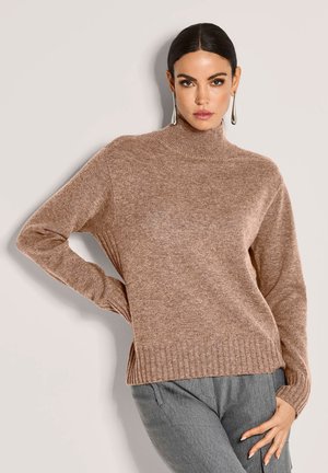 Frau mit nach hinten gekämmtem Haar, die einen langen braunen Strickpullover und graue Hosen trägt und mit einer Hand auf der Hüfte vor einem schlichten Hintergrund posiert.