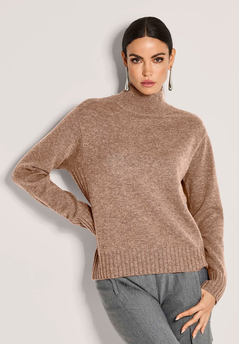 Frau mit nach hinten gekämmtem Haar, die einen langen braunen Strickpullover und graue Hosen trägt und mit einer Hand auf der Hüfte vor einem schlichten Hintergrund posiert.