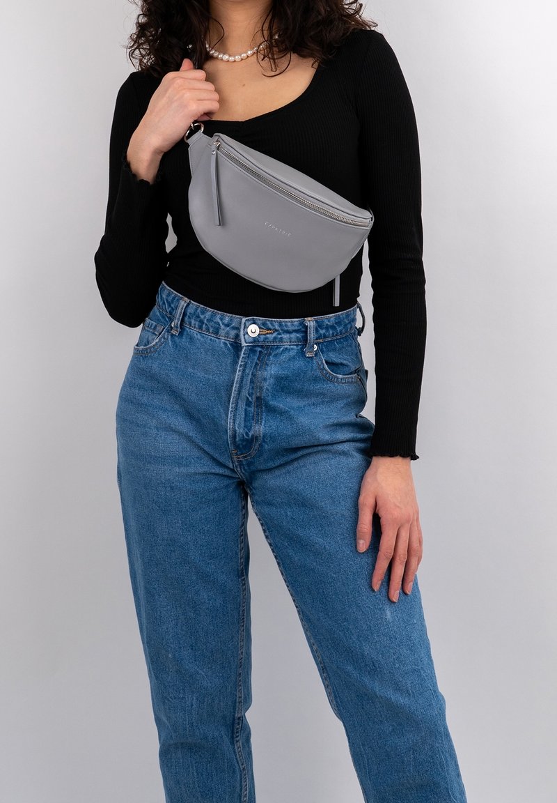 Marsupio in pelle grigia con design curvo, chiusura con zip e zip pull pendente. Abbinato a una maglietta nera a coste e jeans blu.