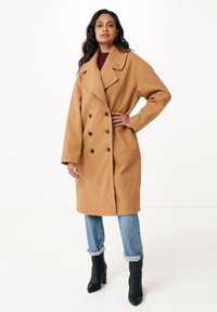 Cappotto camel doppiopetto con grandi bottoni neri, vestibilità over e rever a punta, abbinato a jeans blu con risvolto e stivali neri con tacco alto.