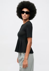 Kobieta z dużą afro nosząca czarną bluzkę peplum z krótkim rękawem, jasnoszare spodnie w prążki i czarne okulary przeciwsłoneczne, ustawiona na tle jasnego jednolitego tła.