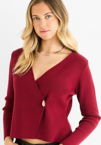 Cardigan rosso lavorato a maglia con scollatura a V, maniche lunghe e chiusura diagonale con bottoni. Presenta un tessuto testurizzato e un design corto.