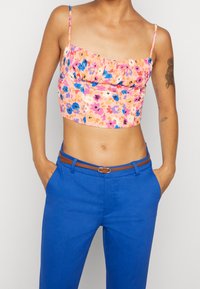Květinový krátký top v pastelově růžové barvě se modrými a fialovými květy, má volánkový výstřih a tenká ramínka, je kombinován s modrými kalhotami a hnědým pásem.