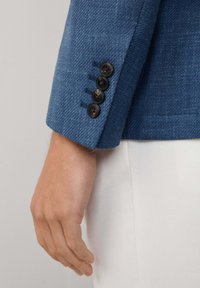 Modrý textured blejzr s rukávem obsahující čtyři černé knoflíky a detailním prošíváním; ruka položená vedle blejzru.