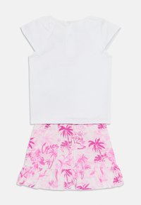 Weiße kurzärmelige Bluse mit geknöpften Rücken, kombiniert mit einem rosa Rock, der mit Palmenmustern und einem voluminösen Saum verziert ist. "Cutes" ist auf dem Rock gestickt.