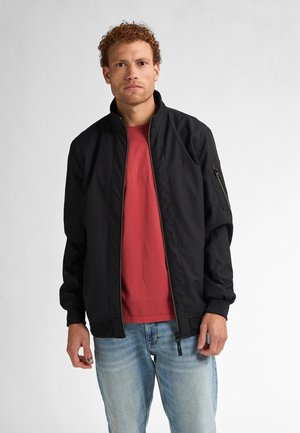 Zwarte bomberjack met een ritssluiting aan de voorkant, geribde manchetten en een zijvak. Draagt over een rood T-shirt en lichtblauwe jeans. Eenvoudig ontwerp.