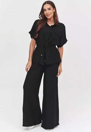 Kobieta ubrana w czarną koszulę z krótkim rękawem zapinaną na guziki z wiązaniem w talii oraz szerokie czarne spodnie, stoi z rękami luźno opuszczonymi wzdłuż ciała.