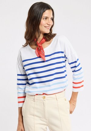 Femme portant un haut blanc avec des rayures bleues et rouges, un pantalon beige, un foulard rouge autour du cou, souriante et regardant sur le côté sur un fond uni.