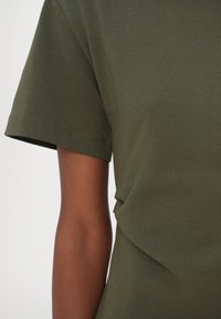 Gros plan d'une personne portant un t-shirt vert olive à manches courtes, montrant la manche et une partie du bras supérieur et du torse.