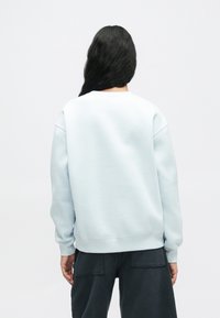 Άτομο με μακριά μαύρα μαλλιά φορώντας ανοιχτό μπλε φούτερ και σκούρα παντελόνια, που στέκεται στραμμένο μακριά σε έναν γυμνό λευκό φόντο.