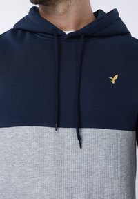 Μπλε σκούρα και γκρι φούτερ με κουκούλα, με υφή στο κάτω μέρος, λείο πάνω μέρος και μικρό χρυσό λογότυπο πουλιού στο στήθος. Περιλαμβάνει κουκούλα με κορδόνι.