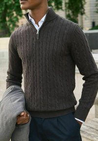 Mörkbrun kabelstickad tröja med halvdragkedja, bärs över en vit skjorta, hålls med en grå kavaj hängande över ena armen.