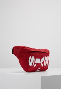 Marsupio rosso in tessuto con chiusura con zip, caratterizzato da un grande testo bianco sulla parte anteriore e una superficie strutturata. Ha una forma curva.