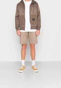 Sweat à fermeture éclair marron avec un panneau texturé et un écusson logo, t-shirt blanc, shorts beiges et baskets beiges avec des accents et lacets blancs.