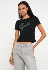 Czarny krótki t-shirt z prążkowanej bawełnianej tkaniny z błyszczącym srebrnym logo "Guess EST.1981" na przodzie. W zestawie z jasnoszarymi dżinsami.