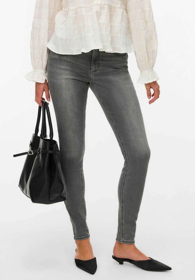 Femme portant un jean gris ajusté, un chemisier blanc à volants, des chaussures slingback noires à bouts pointus, tenant un sac à main en cuir noir.