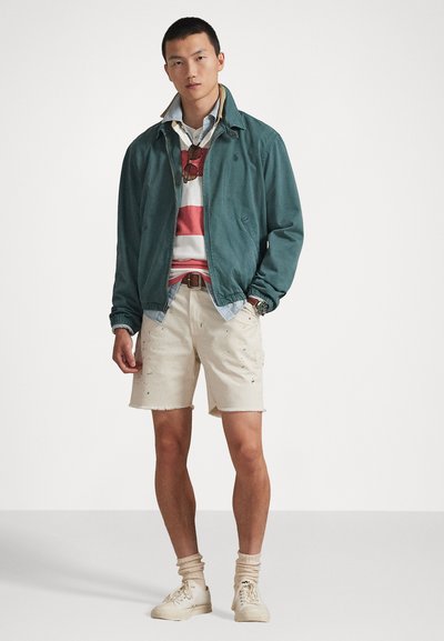 Žalsvai mėlyna striukė ant dryžuotos raudonos ir baltos spalvų marškinėlių, kreminės spalvos nusidėvėjusios šortai, kreminės spalvos kojinės ir sportbačiai, matomi akiniai nuo saulės ir laikrodis.