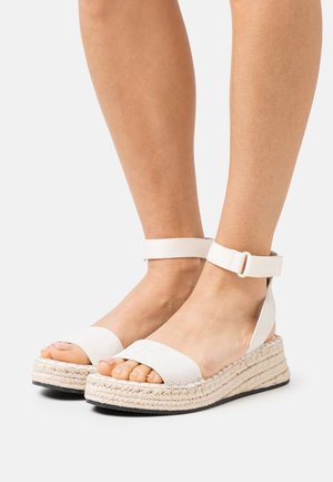 Stopy w białych sandałach na platformie z paskiem na kostce i plecionymi podeszwami stylu słomkowego na jasnym tle.