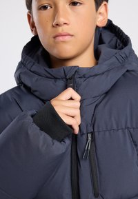 Laivastonsininen puhvijakku hupulla, jossa on joustavat mustat hihansuut ja etuvetoketju. Kankaan pinta on sileä ja siinä on tilkkuompelut.