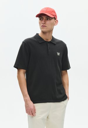 Uomo che indossa una polo nera con logo bianco, pantaloni beige e un berretto rosso, guarda di lato con una mano in tasca su sfondo bianco.