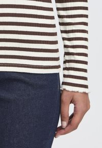 Brun og hvid stribet langærmet top med ribbet tekstur og bølget kant ved manchetterne, parret med mørke denimjeans.