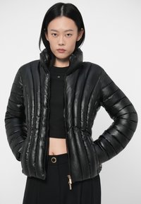 Svart pufferjakke med høy krage, quiltet tekstur og blank finish. Har en frontglidelås og en litt cropped design.