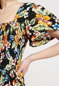 Kwiatowa sukienka na czarnym tle, zdobiona kolorowymi wzorami hibiskusów i liści. Krótkie bufiaste rękawy, teksturowany materiał i dekolt w kształcie kwadratu.