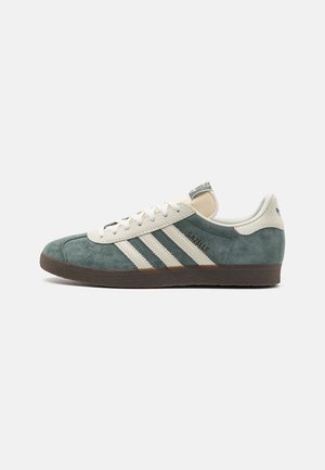 Baskets Adidas Gazelle en daim vert avec des bandes blanches, des lacets beiges et une semelle marron, présentées de profil sur fond blanc.