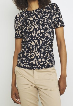 Vrouw draagt een zwart-beige abstract bedrukt topje met korte mouwen en beige broek, staand tegen een effen lichte achtergrond.