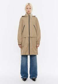 Lichtbeige regenjas met een rits aan de voorkant, verstelbare capuchon en twee zijzakken met rits. Beschikt over elastische manchetten en een relaxed fit. Gecombineerd met brede jeans.