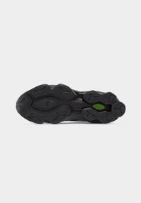 Black athletic shoe sole with textured grip patterns and a small green oval detail near the arch area on a gray background.