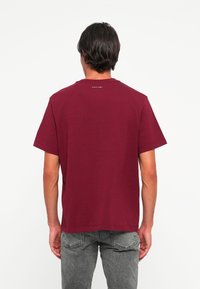 Vīrietis, kurš valkā vienkāršu burgundijas krāsas Calvin Klein t-kreklu, stāv ar muguru pret kameru, kopā ar izbalējušām pelēkām džinsiem.