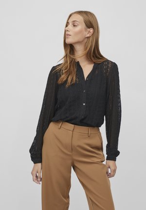 Zwarte kanten blouse met lange mouwen en knoopsluiting, met een gestructureerd patroon en geplooide manchetten, gecombineerd met hooggetailleerde, beige broeken.