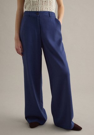 Mujer con pantalones azul marino de talle alto y pierna ancha con bolsillos y zapatos puntiagudos marrón oscuro, combinados con un top sin mangas de crochet color crema.
