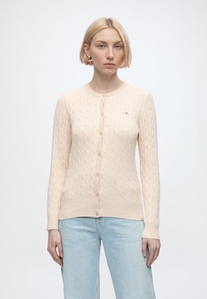 Femme aux cheveux blonds courts portant un cardigan boutonné beige clair en maille torsadée et un jean bleu clair, debout devant un fond blanc uni.