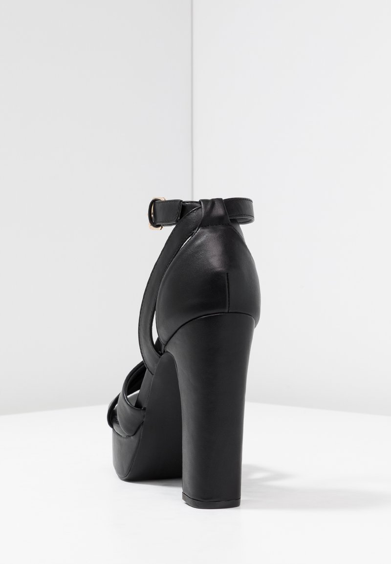 Sandalia de tacón alto de cuero negro con un tacón grueso y correa en el tobillo, que presenta un diseño de punta abierta y textura suave.