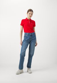 Camisa polo de algodón roja con cuello, combinada con unos vaqueros de denim de tiro alto en azul claro y zapatillas blancas. Diseño simple, sin patrones.