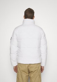 Giacca a vento bianca imbottita con colletto alto. Presenta cuciture orizzontali e una toppa con il logo sulla manica sinistra. Indossata su pantaloni cargo color khaki.