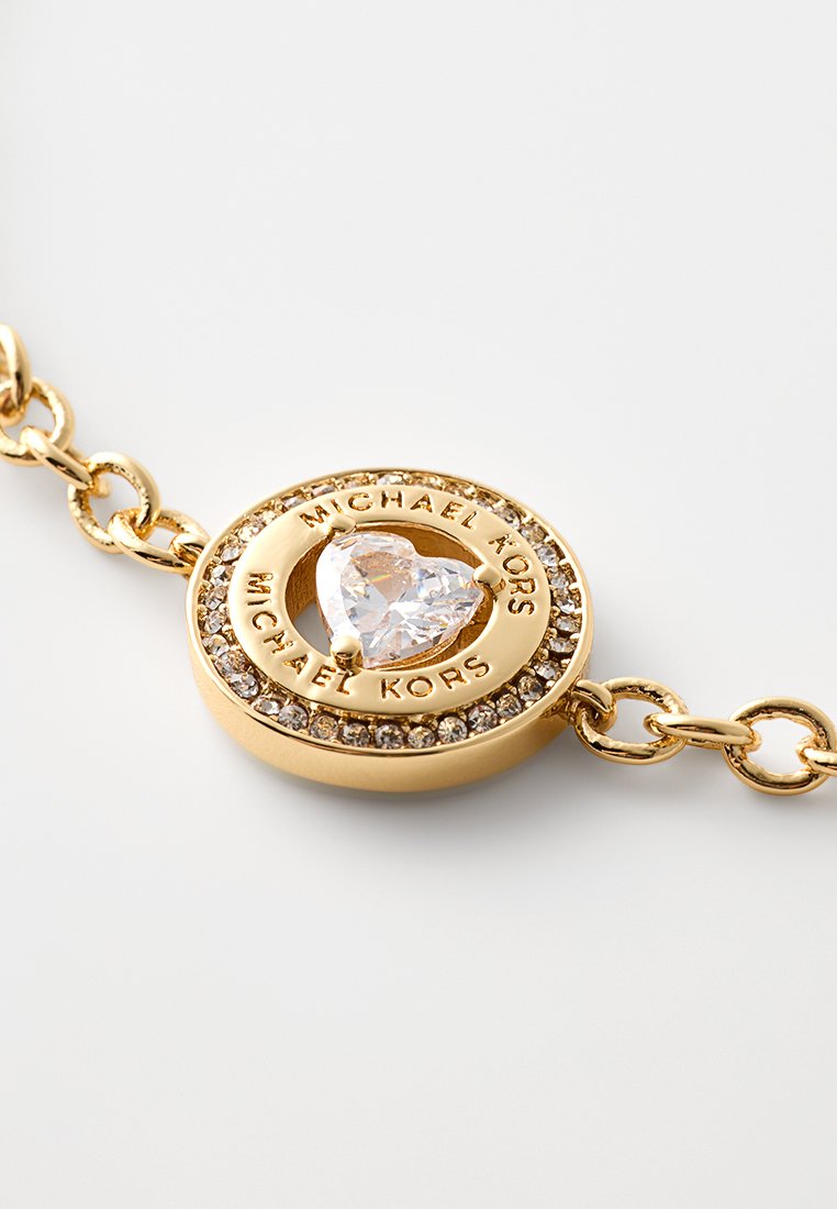 Pulsera en tono dorado que presenta un charm circular con una piedra clara en forma de corazón, rodeada por un anillo de piedras más pequeñas y un logo grabado.