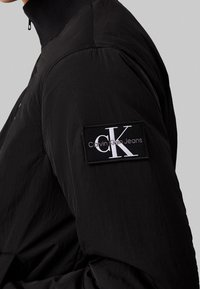 Svart jackärm med en svart lapp som har vit text "CK" och "Calvin Klein Jeans" på överarmen mot en neutral bakgrund.