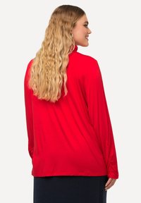 Rode longsleeve top met een hoge hals. Soepele textuur, relaxte pasvorm en plooien bij de mouwen. Draag het met een donkerblauwe rok.