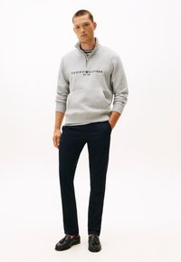 Grijze fleece pullover met een kwart ritssluiting, met een logo op de borst, gecombineerd met marineblauwe broek en zwarte loafers.