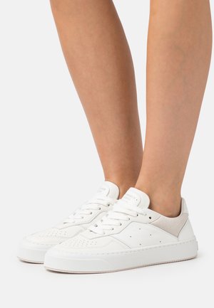 Legs wearing white low-top leather sneakers with laces and perforated details, standing against a plain light background.