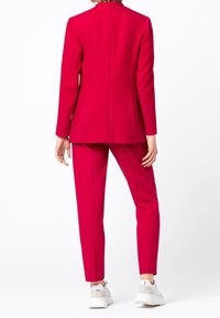 Completo rosso con blazer su misura e pantaloni slim. Realizzato in tessuto liscio, presenta un design pulito. Abbinato a sneakers bianche.