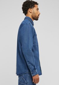 Denim-paita, jossa on nappikaulus, pitkät hihat, rintatasku ja normaali mitoitus keski-sinisessä värissä. Tasainen rakenne ja klassinen muotoilu.