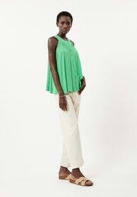 Groene mouwloze top met knoopdetails, gecombineerd met beige wijde broek en bruine wedge sandalen met kruisbanden. Glad stofoppervlak.