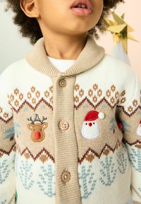Kremų ir smėlio spalvos kardiganas su apsiaustu, papuoštas siuvinėtų elnių ir Kalėdų Senelio motyvais, rudais mygtukais ir žiemos rašto dizainu.