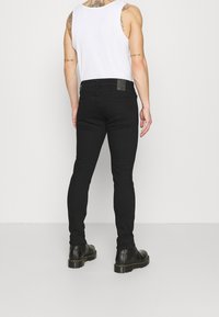 Jeans negros ajustados con una etiqueta de cuero en la cinturilla trasera, combinados con una camiseta sin mangas blanca y botas de tobillo negras, vistos desde la parte de atrás.