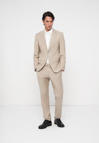 Férfi áll fehér háttér előtt, bézs öltönyt, fehér inget és fekete elegáns cipőt viselve, kezével a zsebében.