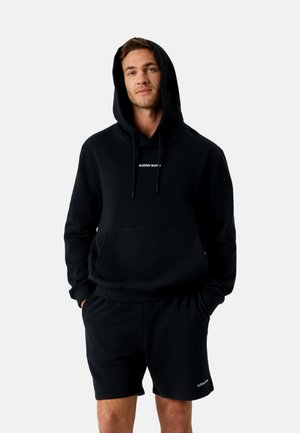 Férfi fekete kapucnis pulóverben és hozzáillő rövidnadrágban, elöl zsebekkel, kezeit zsebekben tartva, egyszínű háttér előtt áll.